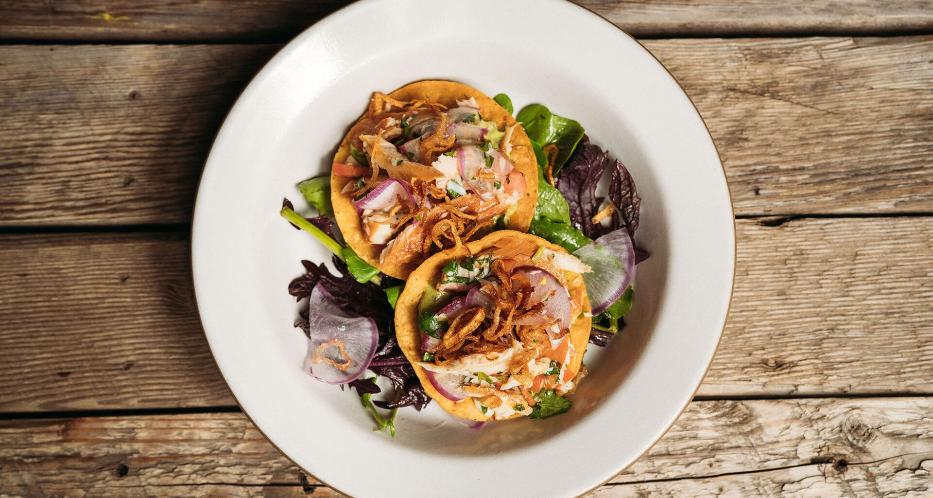 Overhead view of a plate of two tostadas topped with fish and fried shallots, served on a wooden picnic table.