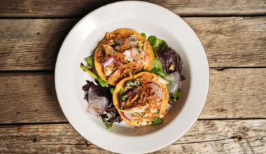 Overhead view of a plate of two tostadas topped with fish and fried shallots, served on a wooden picnic table.