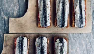 Six rectangular toasts topped with whole sardines, arranged on two wooden cutting boards against a gray marble surface.