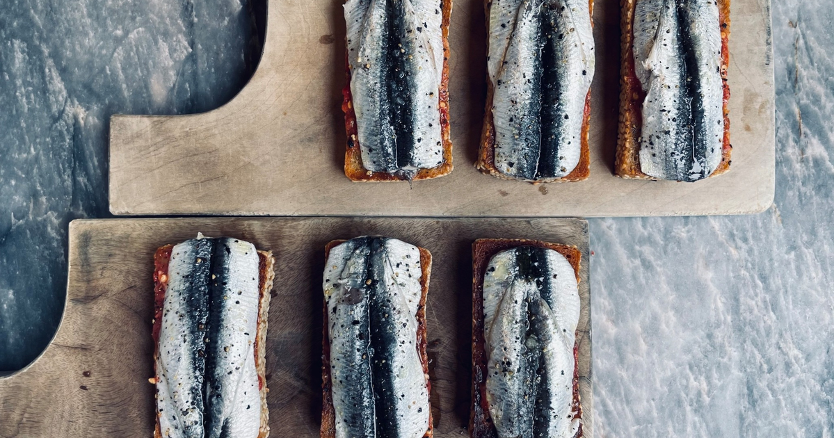 Six rectangular toasts topped with whole sardines, arranged on two wooden cutting boards against a gray marble surface.