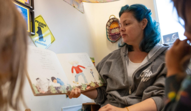 A woman with blue hair reads a picture book aloud to children in a cozy room with colorful decorations and toys.