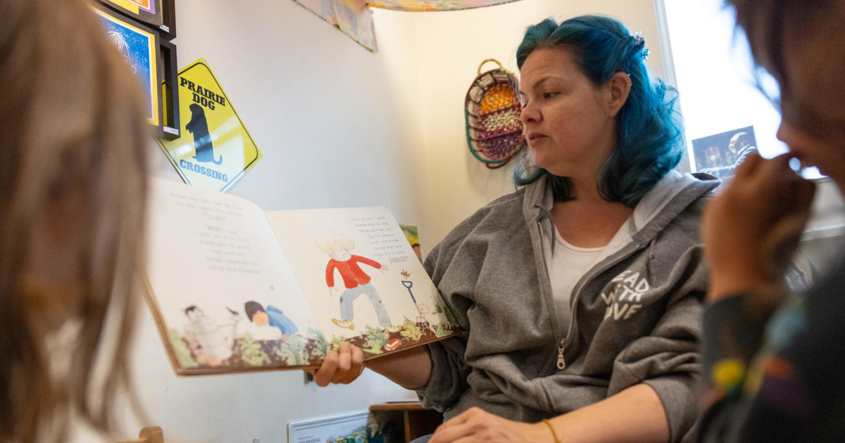 A woman with blue hair reads a picture book aloud to children in a cozy room with colorful decorations and toys.