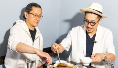 A man wearing round glasses and a beige hat eats dumplings with chopsticks, holding a spoon under the food, seated at a table with bowls of soup and dishes.