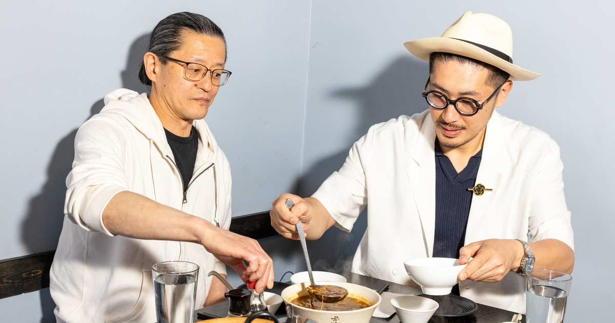 A man wearing round glasses and a beige hat eats dumplings with chopsticks, holding a spoon under the food, seated at a table with bowls of soup and dishes.