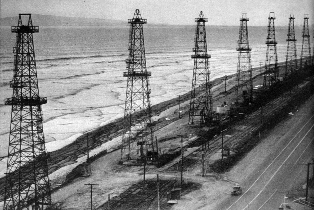 A black and white image showing multiple oil drilling platforms and towers along a beach