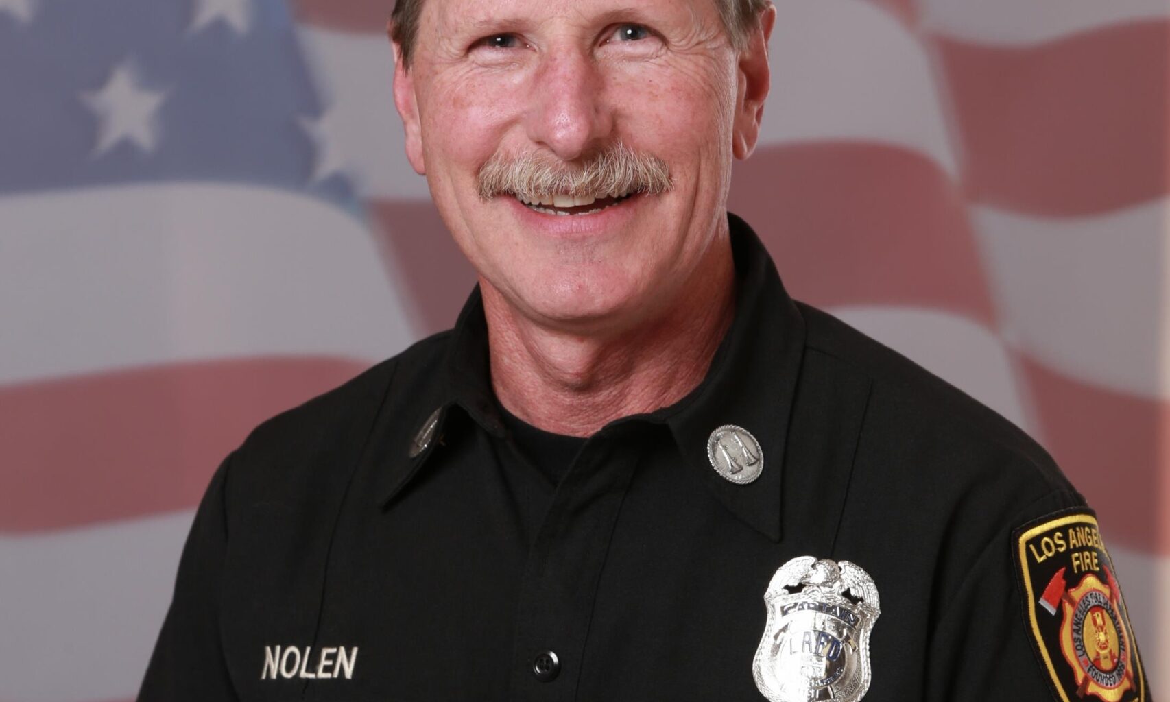 Portrait of a smiling man in a black firefighter uniform with patches, set against a blurred American flag background, conveying pride and professionalism.