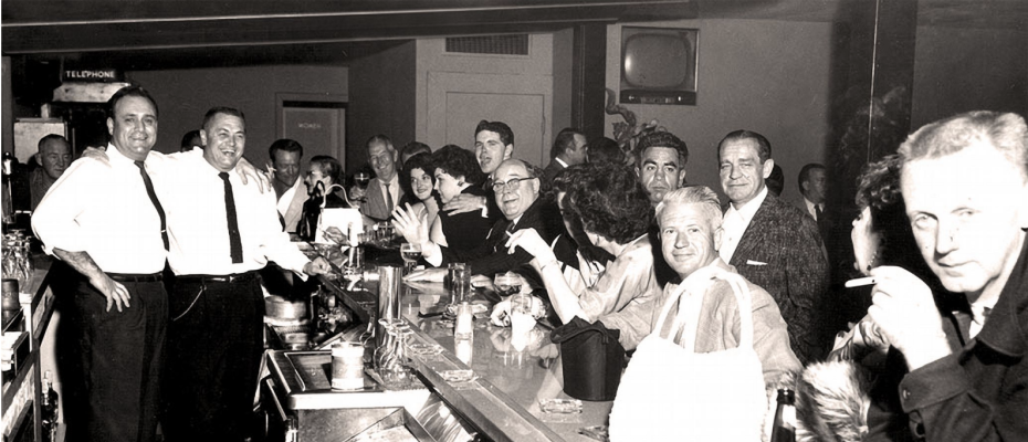 Black and white photo of a crowded bar, with people sitting and standing at the counter, some smiling, some smoking, and bartenders serving drinks.
