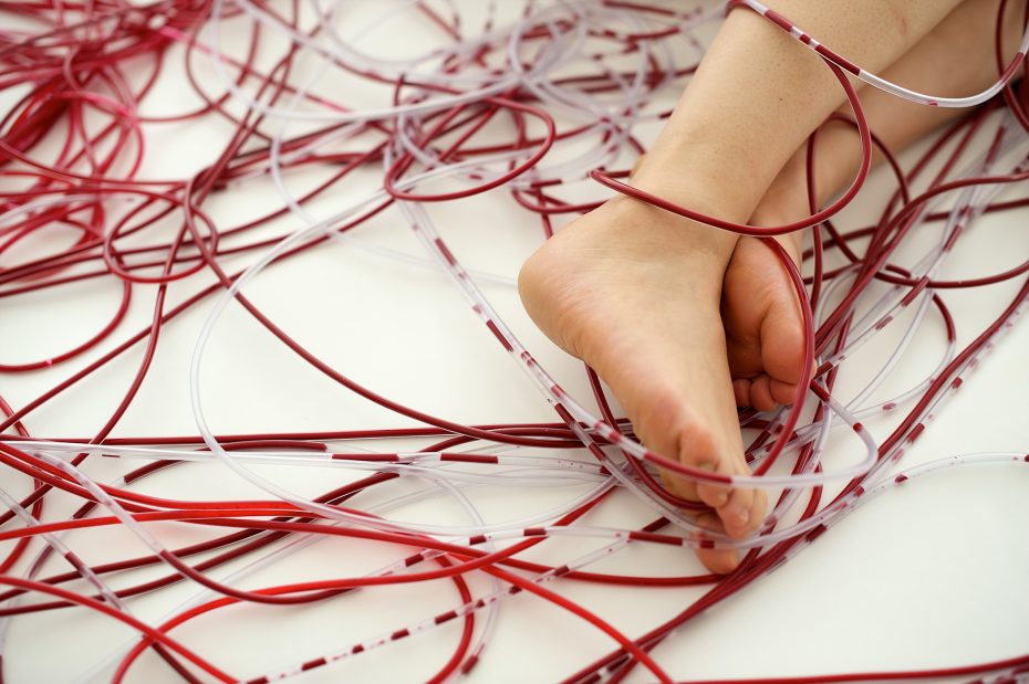 Bare feet resting on a white surface surrounded by tangled red and clear plastic tubes.