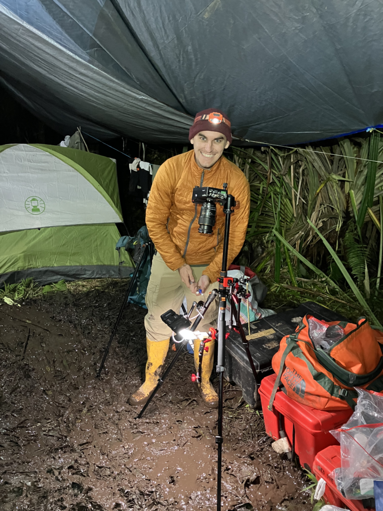 a smiling man wearing a head lamp standing in a mud puddle under a tarp as he sets up a camera and lights