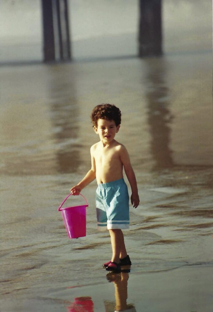 A young, shirtless David holding a pale and standing near the tideline at the beach