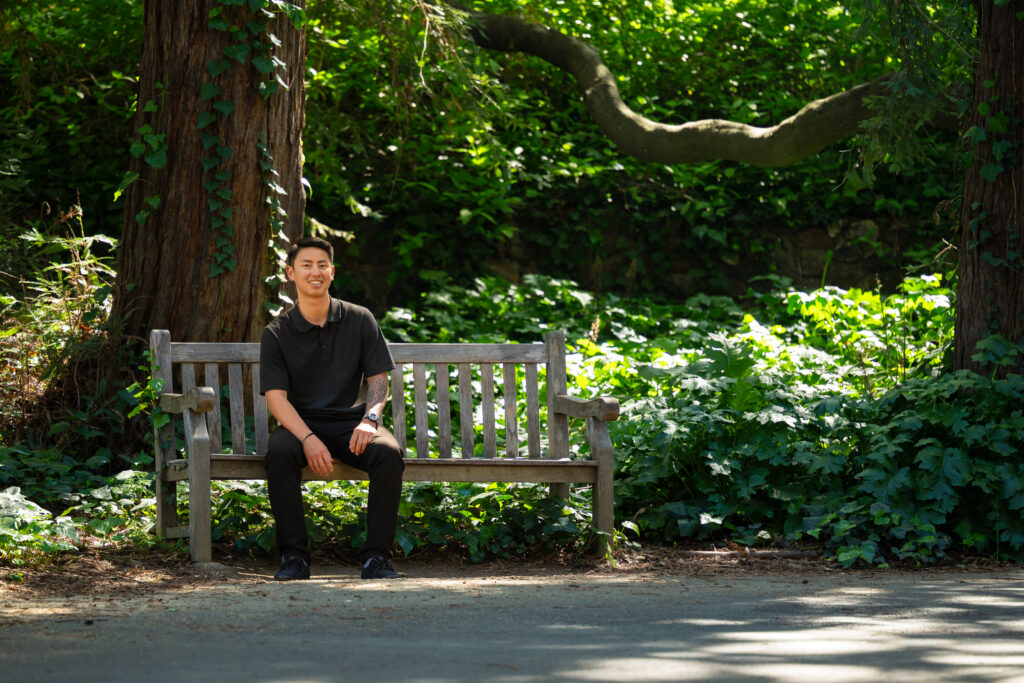 Dean Tanioka sits on a bench on campus, near Strawberry Creek, surrounded by trees and greenery