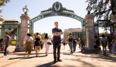 Dean Tanioka stands in front of Sather Gate, with his arms crossed, while students who are slightly blurred in the background walk by.