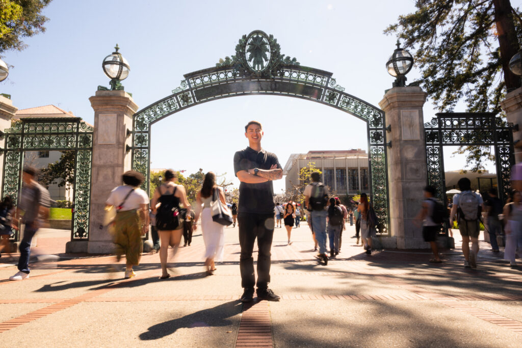 Dean Tanioka stands in front of Sather Gate, with his arms crossed, while students who are slightly blurred in the background walk by.