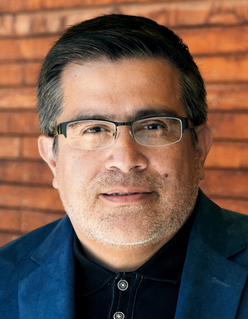 Casual headshot of UC Berkeley economist Enrique Lopezlira, wearing a black shirt and a blue jacket, standing again a wall of rust-red bricks