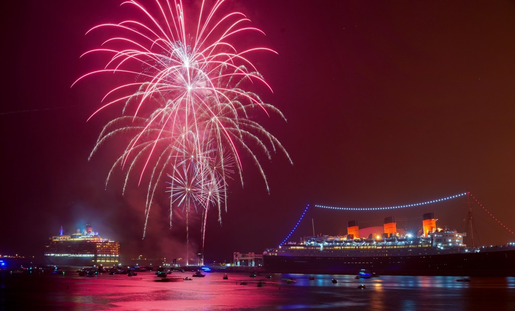 Fireworks explode over a large ship and smaller boats in the water at night.