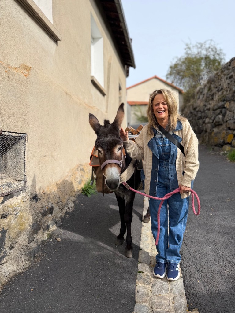Woman standing next to a donkey on a narrow path beside a beige building.
