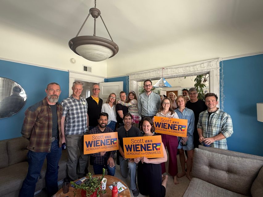 A group of people standing and sitting in a living room, some holding orange signs that read "Scott Wiener Democrat for Congress.