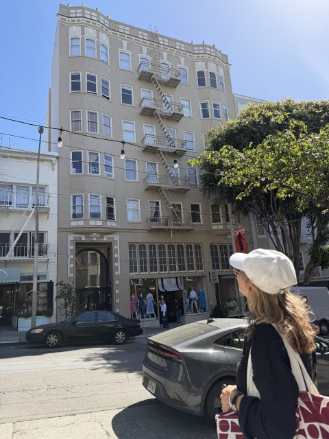 A woman in a white cap stands on a city sidewalk, looking at a beige six-story building with fire escapes and storefronts at street level. Cars are parked along the curb.