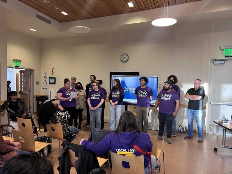 About a dozen people, many of them in matching purple shirts, stand at the front of a room while one person reads from a sheet of paper.