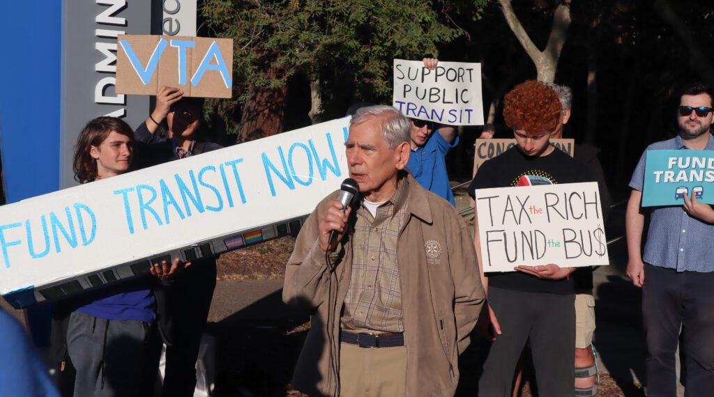 Photo of a crowd of people standing outside, holding signs reading "Fund transit now" and "Tax the rich, fund the bus" and "Support public transit." The photo is centered on Rod Diridon, an elderly man with white hair wearing a beige jacket with the Rotary Club logo