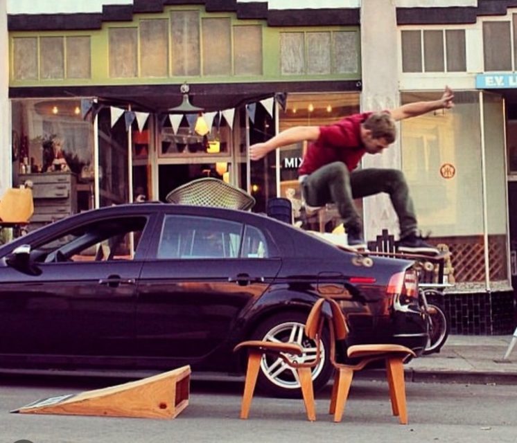 A person on a skateboard jumps over a chair placed on top of a black car, with a shopfront in the background.