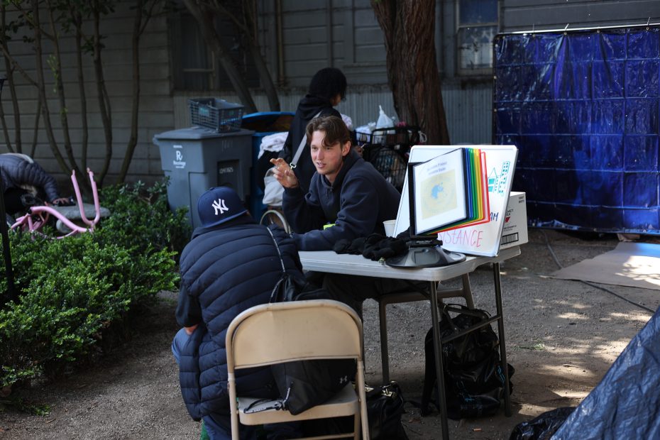 Two people sit across from each other at an outdoor table, having a conversation. Papers, a plastic display, and gloves are on the table. Others and various items are in the background.