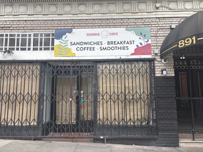 A storefront with a closed security gate and a sign reading "Sammie Cafe - Sandwiches, Breakfast, Coffee, Smoothies." Windows are covered with brown paper.