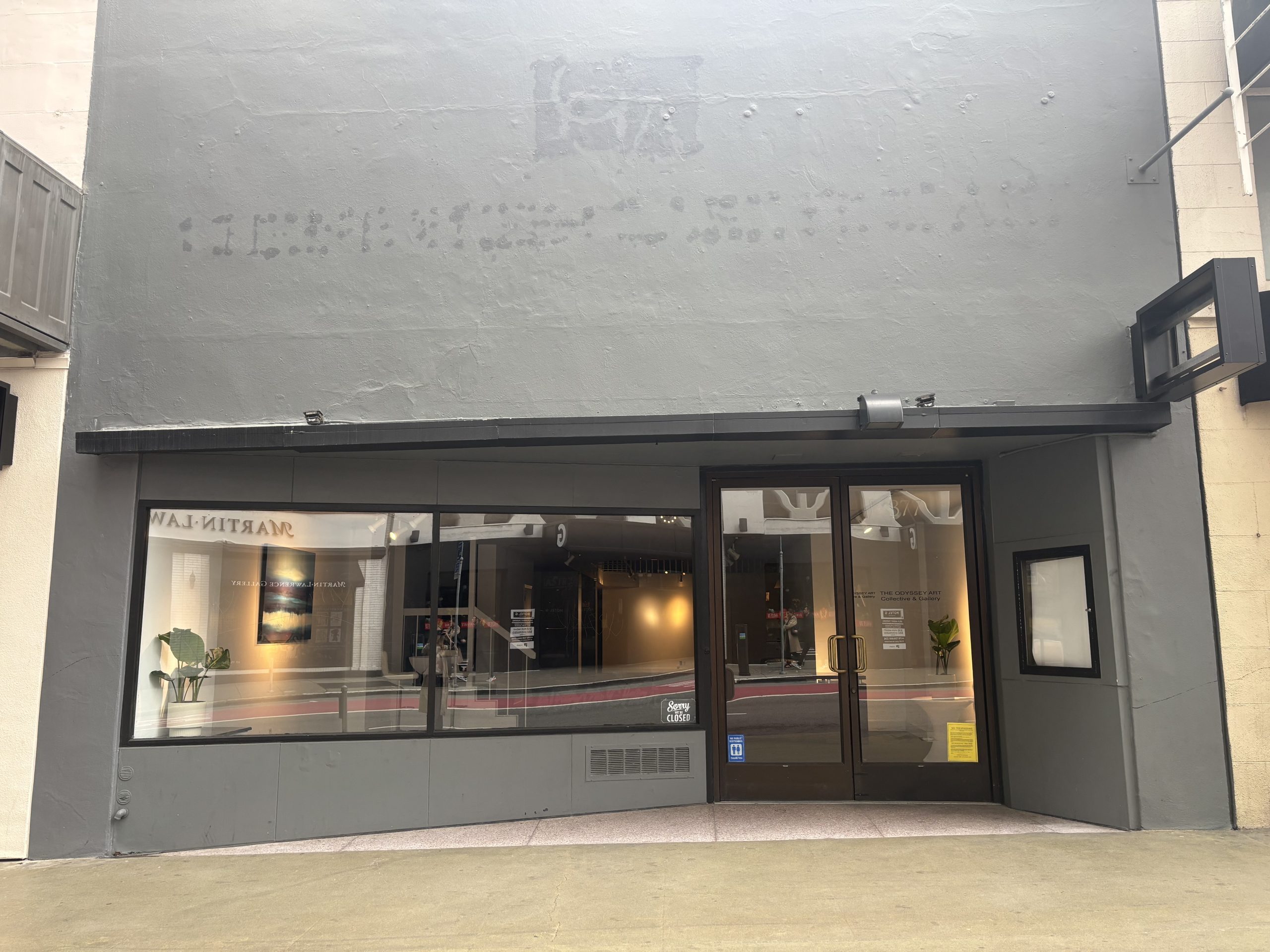 A gray storefront with large windows, glass double doors, and minimal signage. The interior is visible, featuring plants and simple decor.