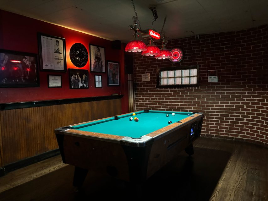 A pool table with balls set up in a dimly lit bar, featuring red lamps, framed photos on red and brick walls, and a wooden floor.