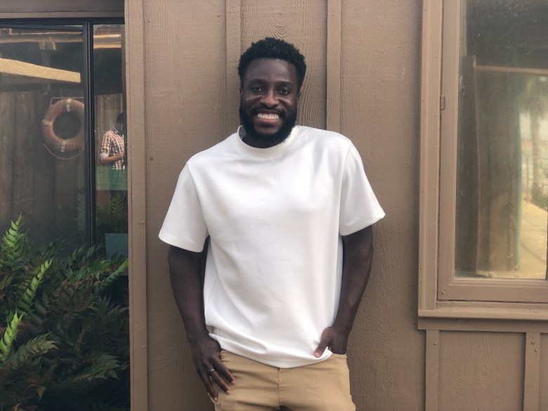 A smiling Black man with a beard, wearing a white T-shirt and khaki pants, standing against a brown wall.