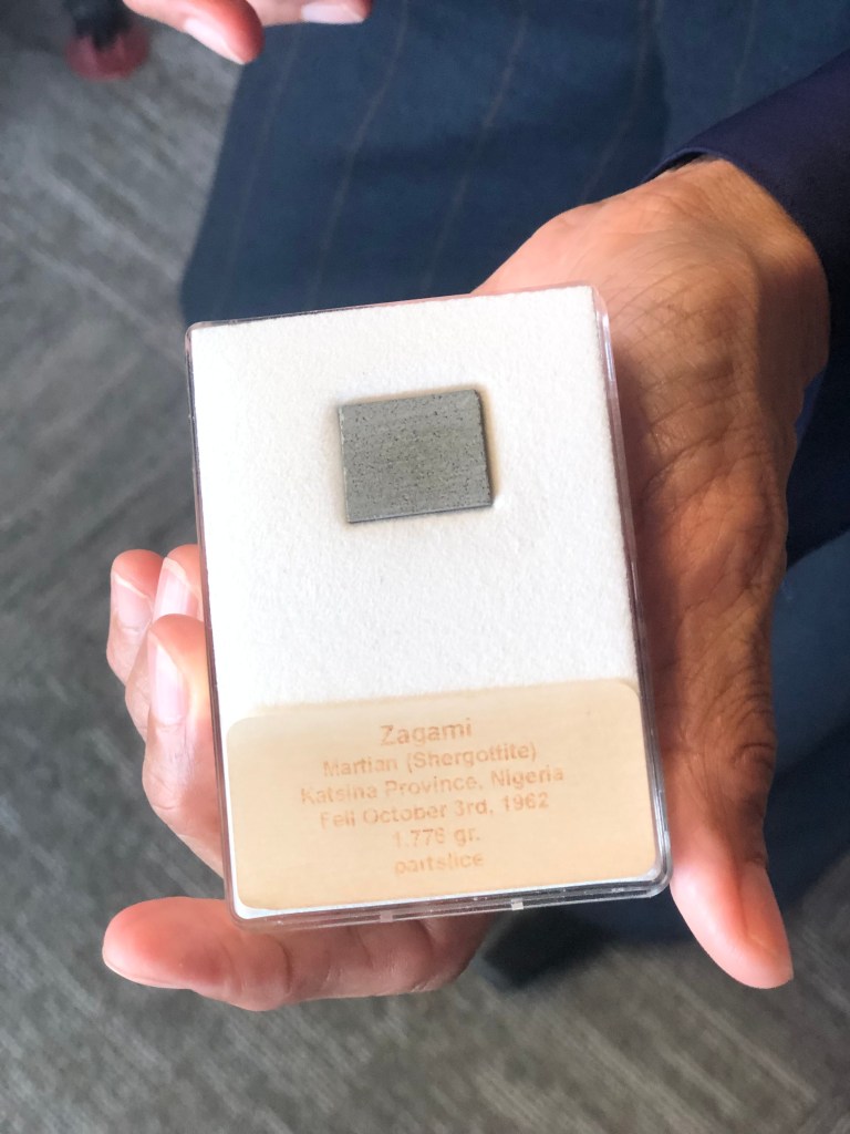 A small piece of grey rock, actually a meteorite, in a box that says: "Zagami, Martian (Shergottite), Katsina Province, Nigeria. Fell October 3rd, 1962. 1,775 gr. parts/ice."