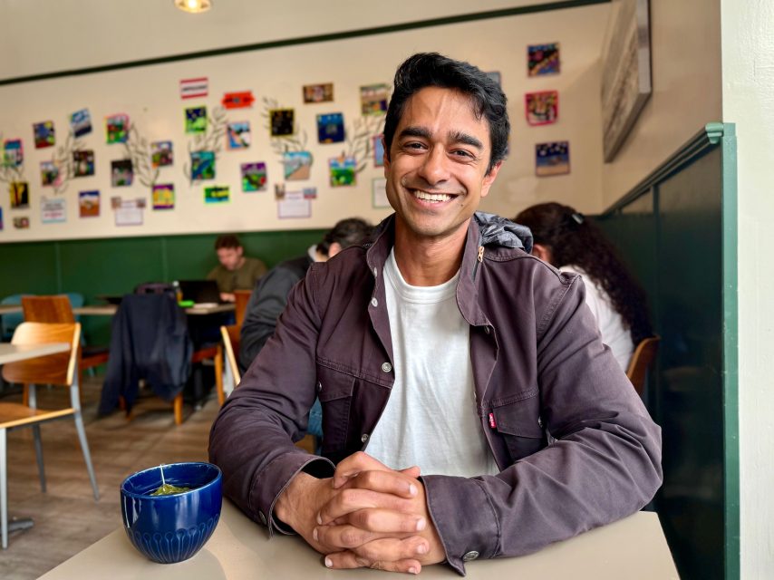 A man sits at a table in a café, smiling at the camera with his hands folded on the table. A blue mug is in front of him; people and artwork are visible in the background.