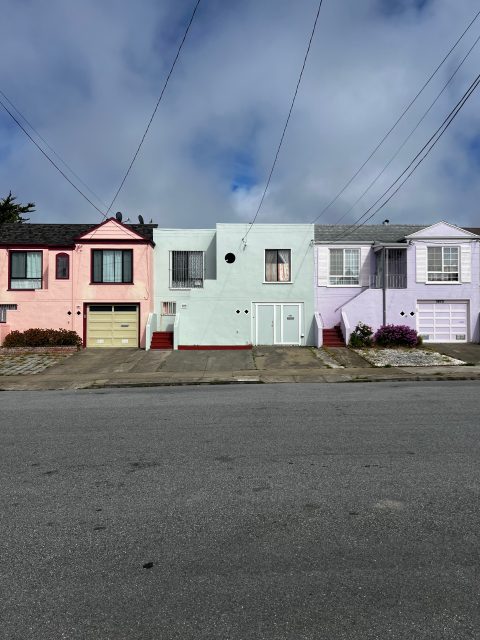Three pastel-colored houses—pink, light blue, and lavender—stand side by side on Noriega Street with steps leading to their entrances, set against a cloudy sky and an empty street in the foreground.