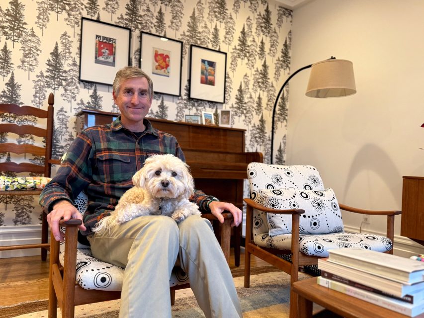 A man sits on a patterned chair in a living room with a small white dog on his lap. There are pictures on the wall, a lamp, a piano, and a stack of books on a table.