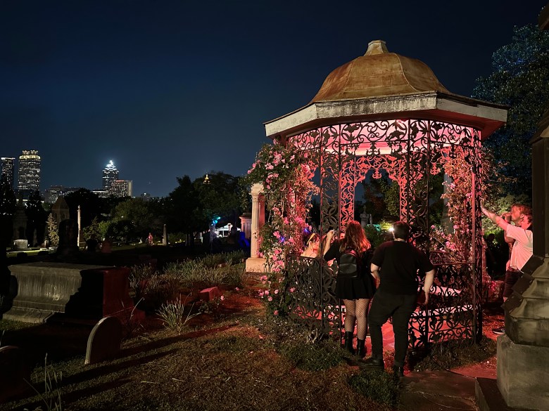 Visitors at rose-covered iron gazebo glowing pink during illumine 2025 at Oakland Cemetery Atlanta, skyline in background