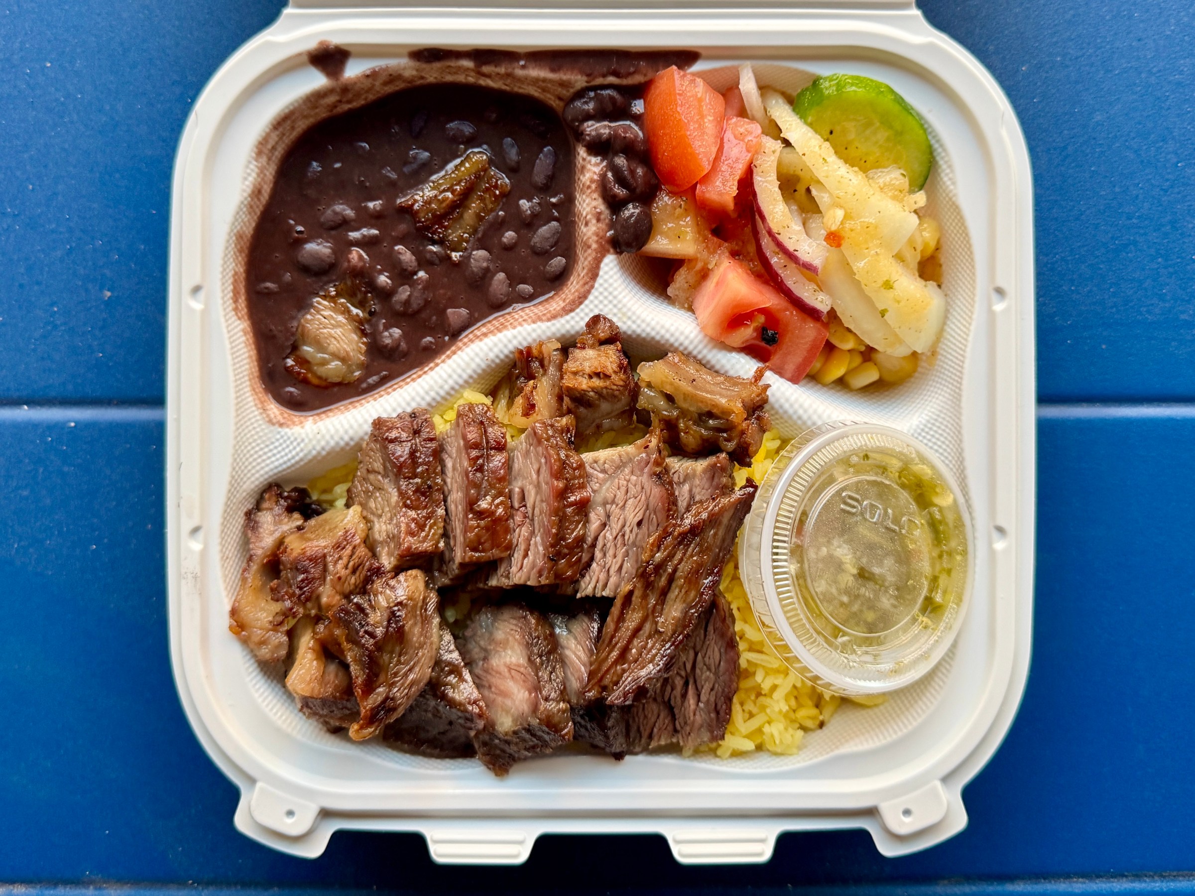 A plastic takeout box filled with beans, salad, and rice topped with asado, pork belly, and sausage.