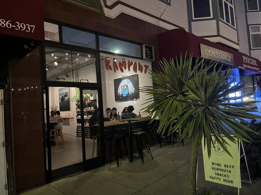 Nighttime street view of Rampant, a bar with people sitting inside and outside, a sign advertising wine, beer, vermouth, snacks, and happy hour, and a palm plant by the entrance.