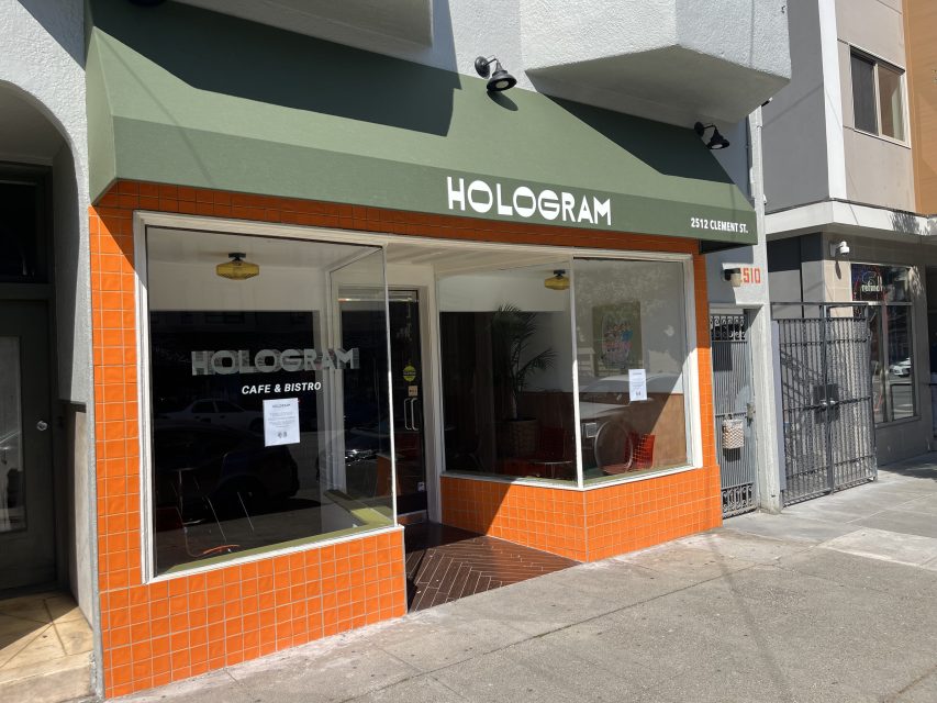 Street view of Hologram Café & Bistro with green awning, orange-tiled exterior, and large front windows on a sunny day.