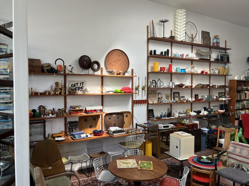 A room with mid-century modern furniture, several shelves filled with vintage objects, books, art pieces, and various collectibles.