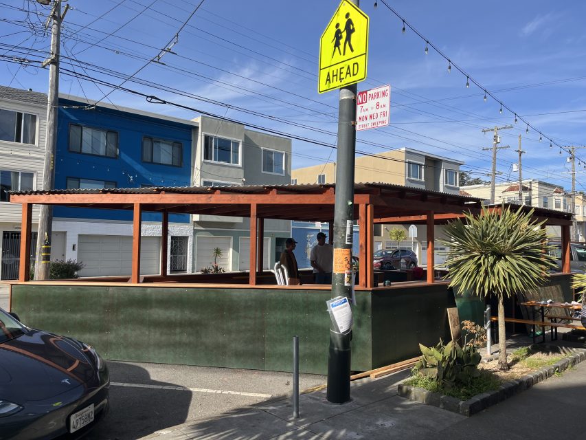 Outdoor dining structure with a corrugated roof and green walls on a street, with a school crossing sign and residential buildings in the background.