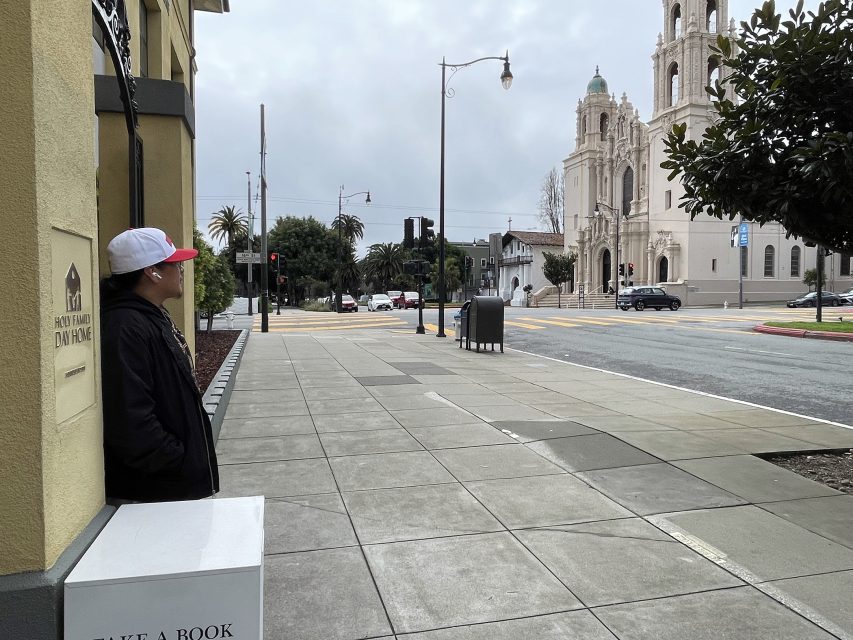 A person in a black jacket and cap stands on a sidewalk near a “Take a Book” box, looking across the street toward a large church building.