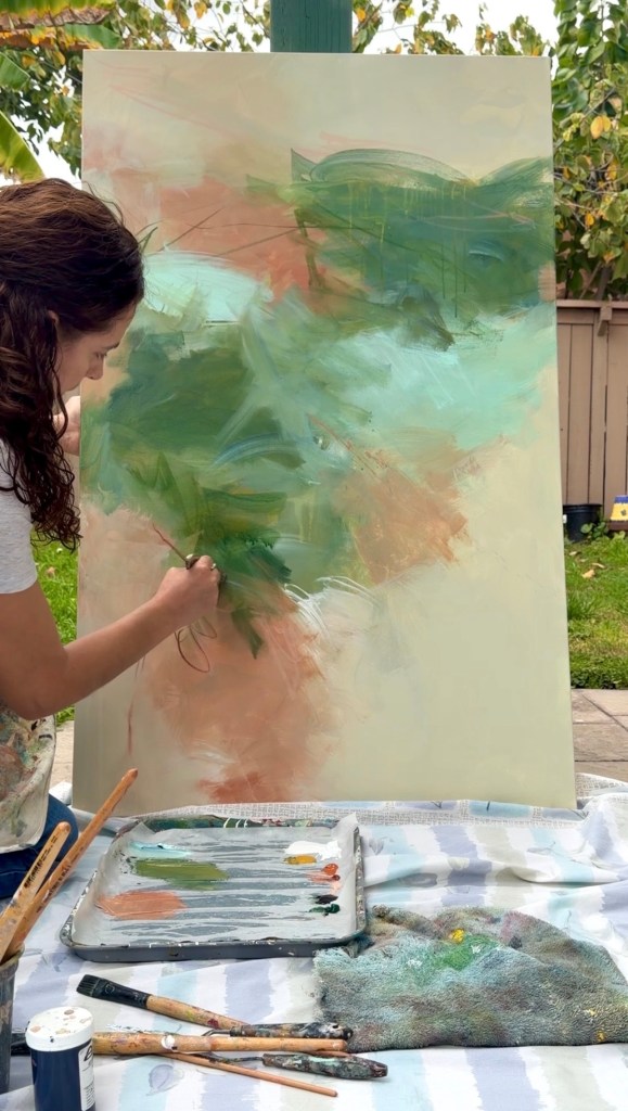 Person painting an abstract artwork on a canvas outdoors, with a palette and paintbrushes on a nearby table.