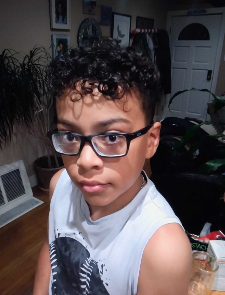 A photo of a child wearing glasses and looking into the camera as they stand in the middle of a living room.