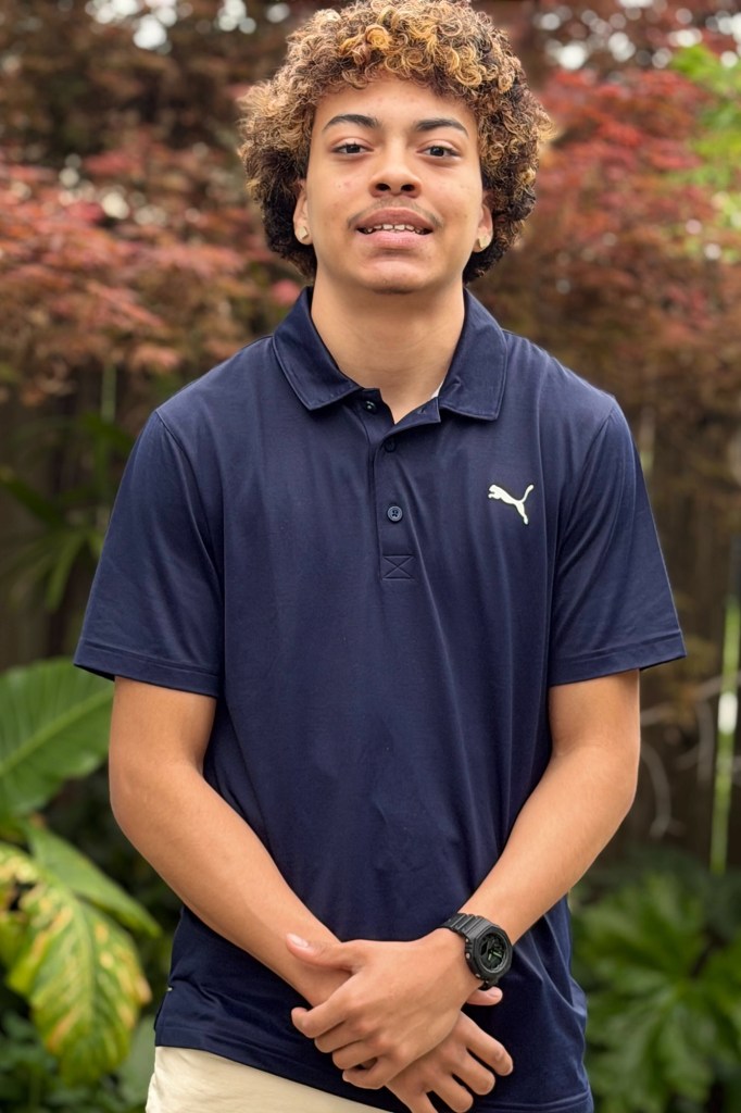 A teenager, wearing a blue shirt and khaki pants, rests their hands in front of them as they pose for a photo.