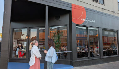 Two women stand talking outside a restaurant with large windows and a red sign that reads "kitchen Istanbul." Diners are visible inside the restaurant.