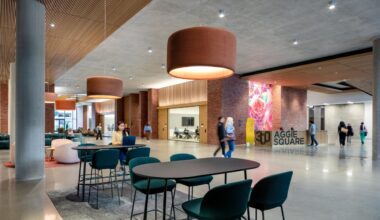 Modern lobby with light fixtures, tables and seating areas, and people interacting. A reception desk has a "300 Aggie Square" banner on it.
