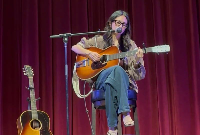 NIKI plays an acoustic guitar while singing into a microphone onstage.
