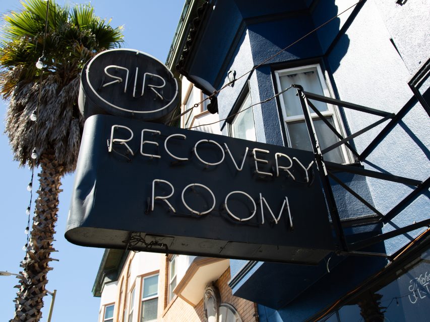 Black neon sign reading "Recovery Room" attached to a blue building, with a palm tree and part of another building visible in the background.