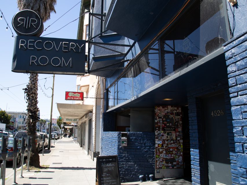 Street view of the exterior of Recovery Room bar with a sign, sidewalk chalkboard, and nearby buildings visible under daylight.