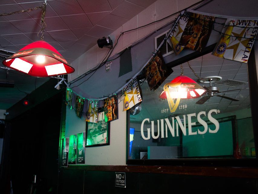Bar interior with a Guinness logo on a glass panel, red hanging lights, wall posters, and a string of decorative flags above.